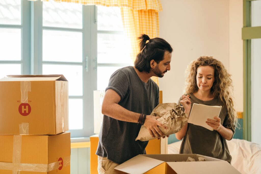pexels-photo-4247768-1 Cheerful young man and woman smiling while unpacking carton boxes with belongings in new apartment during relocation and looking at paper