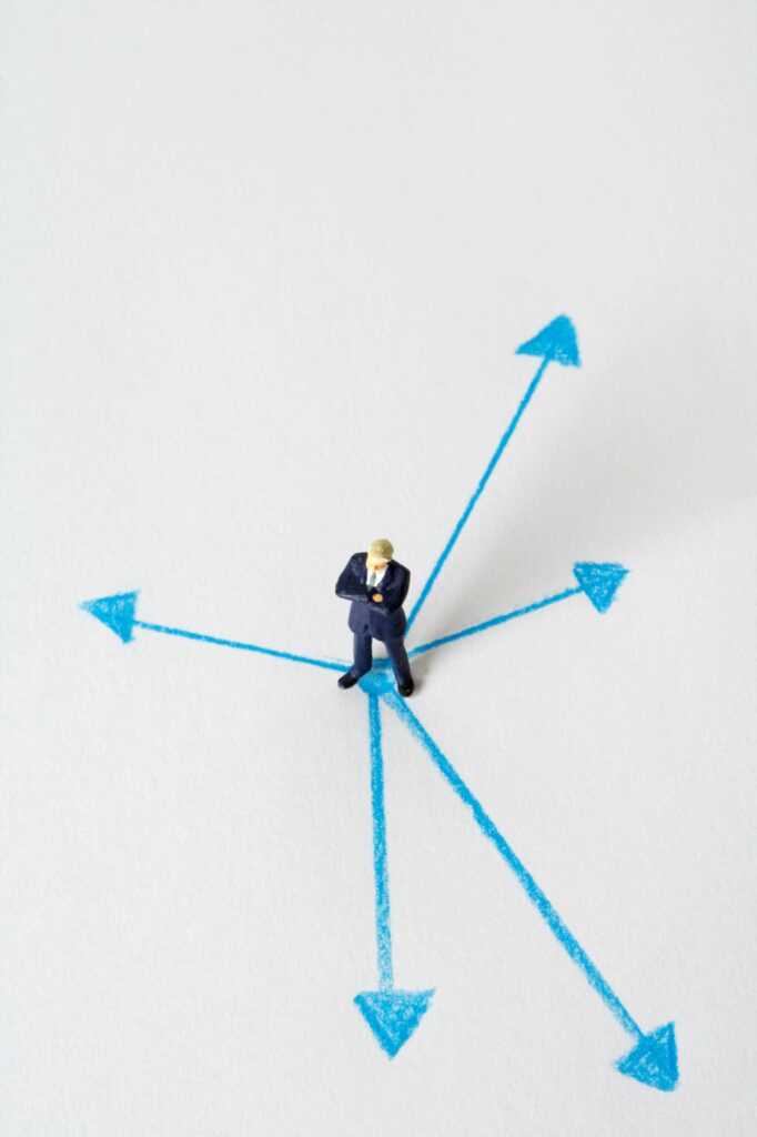 pexels-photo-12585521-12585521 Miniature businessman stands at crossroads of blue arrows on white background, symbolizing choices.